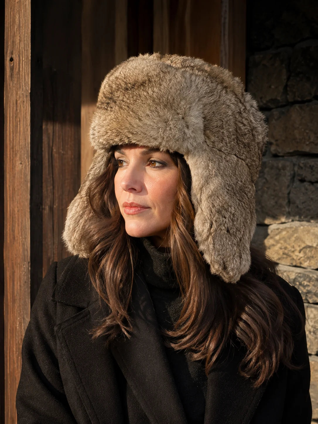 Woman wearing a taupe rabbit fur trapper hat beside a rustic winter cabin