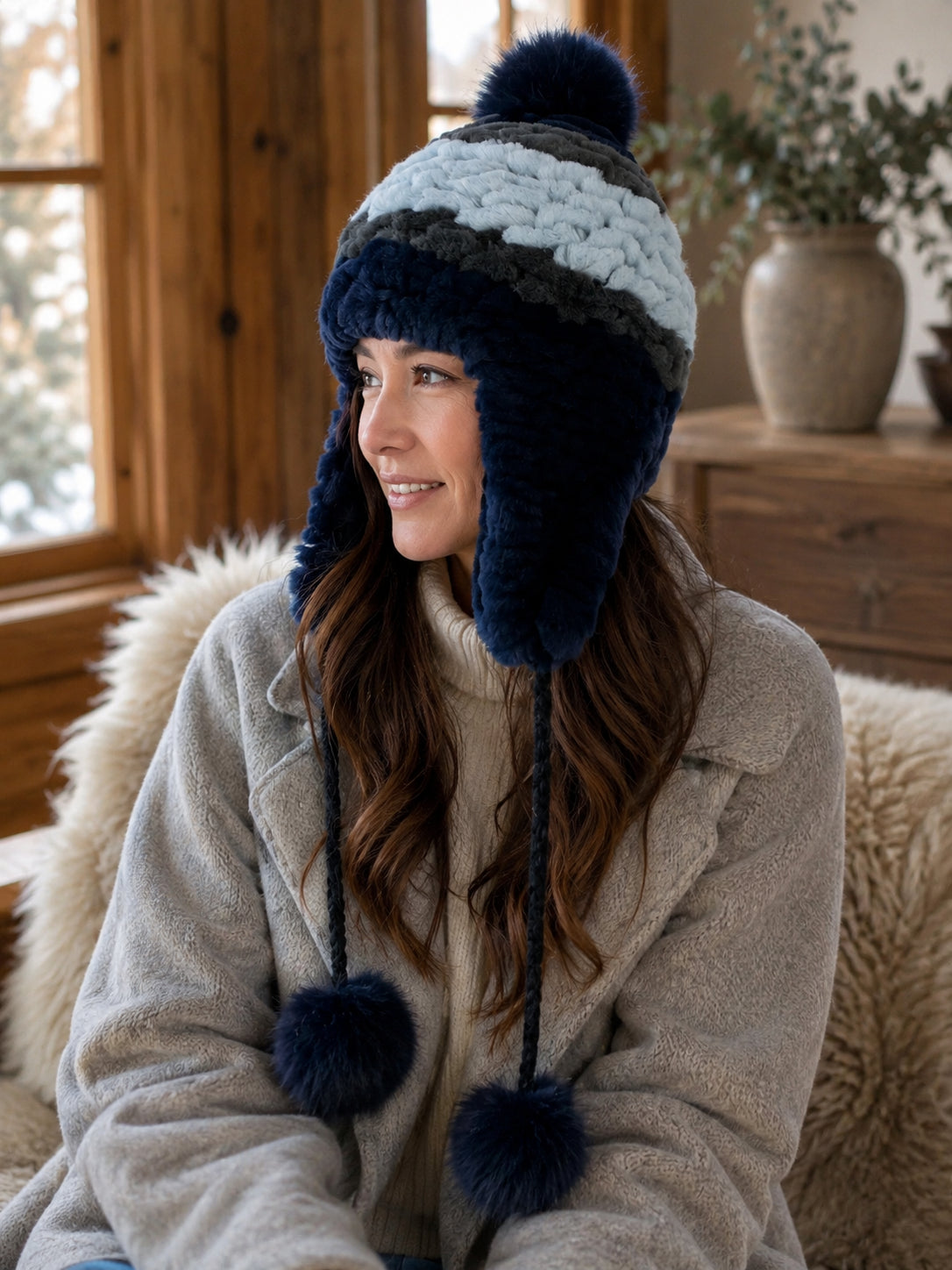 Women's blue Rex rabbit fur earflap hat with fox fur pom-poms in a quiet winter interior