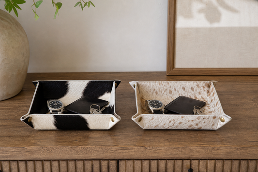 Black and ivory and speckled tan cowhide valet trays compared at the same size on warm wood