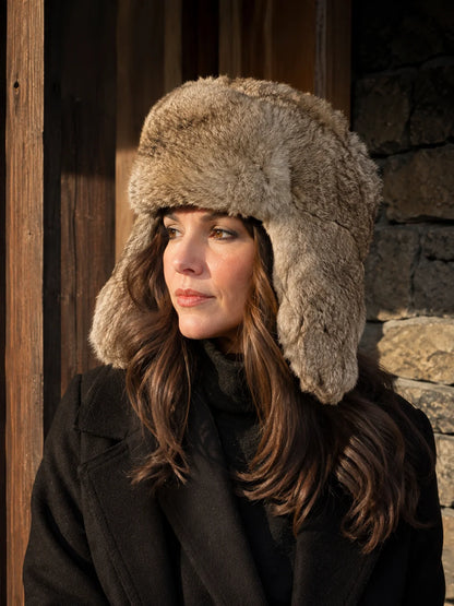 Woman wearing a taupe rabbit fur trapper hat beside a rustic winter cabin
