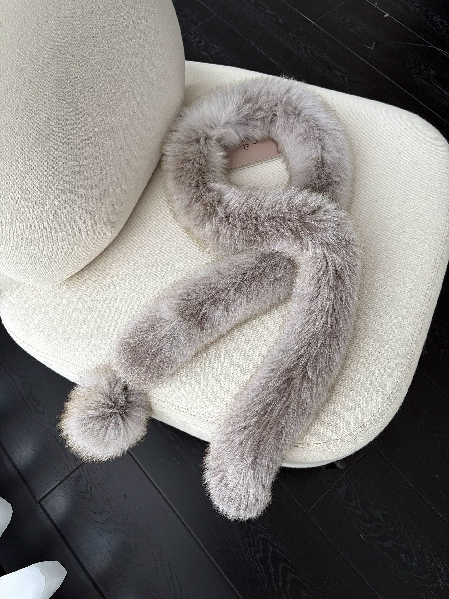Taupe Gray fox fur long scarf with pom-pom arranged on a cream chair