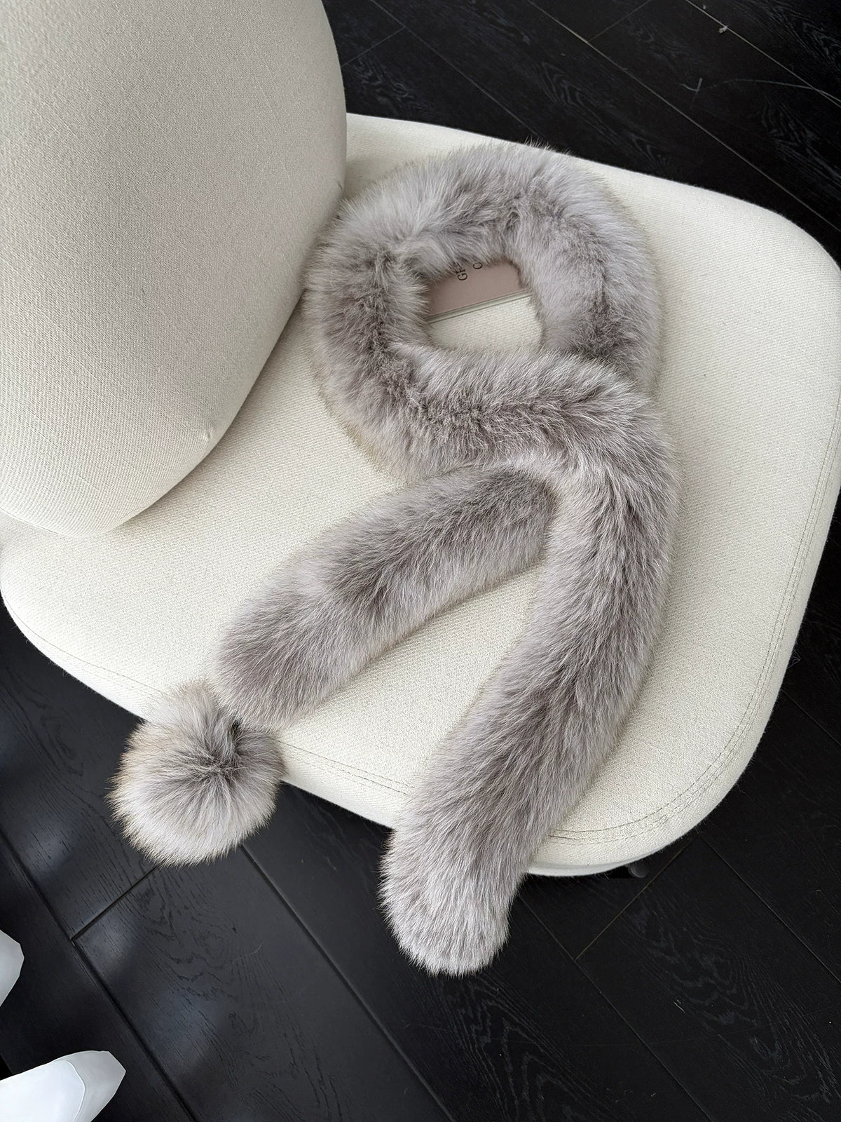 Taupe Gray fox fur long scarf with pom-pom arranged on a cream chair