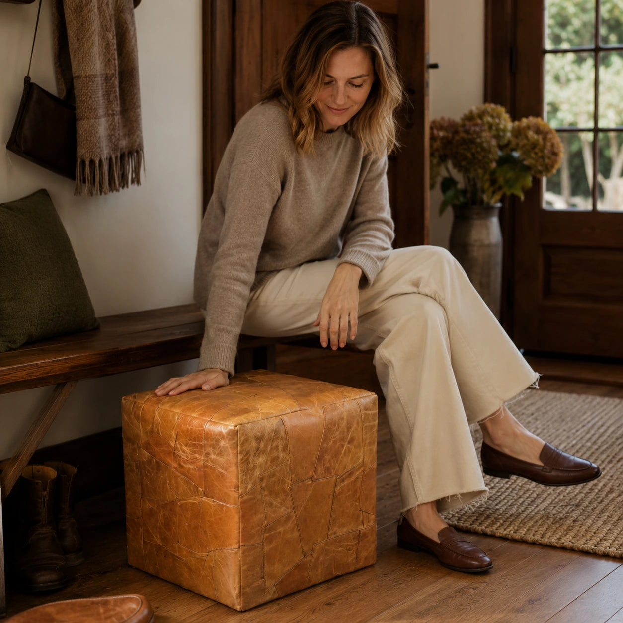 Woman over 35 seated on a honey brown leather cube ottoman in an entryway