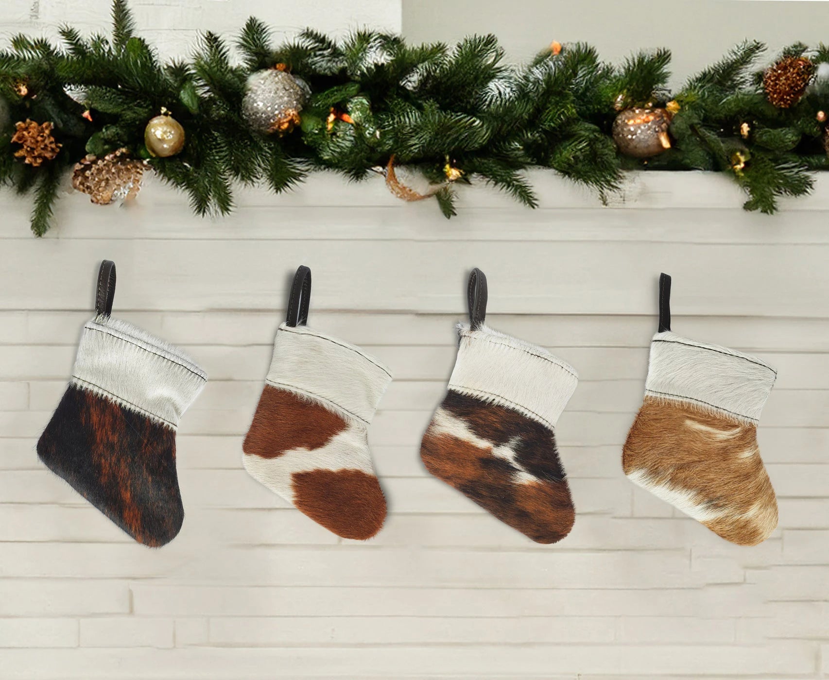 Capricornus Living Four-piece cowhide mini holiday stocking ornament set displayed beneath an evergreen mantel garland