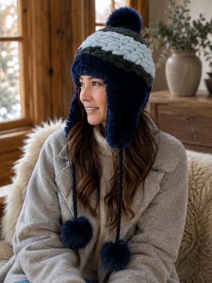 Women's blue Rex rabbit fur earflap hat with fox fur pom-poms in a quiet winter interior